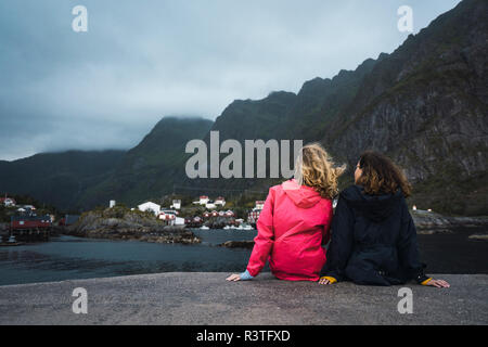 Norvegia Lofoten, vista posteriore di due giovani donne seduto su di un molo presso la costa Foto Stock