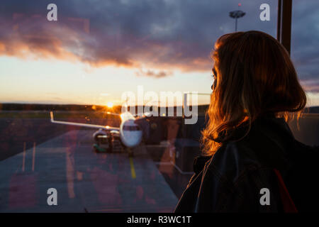 Giovane donna guardando attraverso la finestrella sul piano dell'aeroporto al tramonto Foto Stock