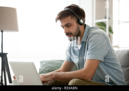 Focalizzato uomo millenario in cuffia utilizzando computer online Foto Stock
