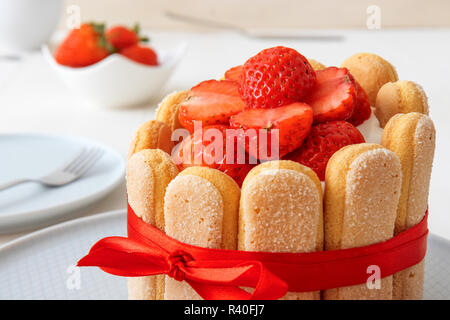 Charlotte, francese dessert con fragole, legato con un nastro rosso scarlatto, tazza di caffè, la ciotola con le fragole sul tavolo bianco. Foto Stock