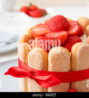 Charlotte, francese dessert con fragole, legato con un nastro rosso scarlatto, tazza di caffè, la ciotola con le fragole sul tavolo bianco. Foto Stock