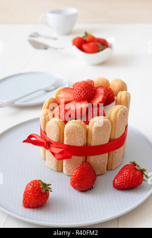 Charlotte, francese dessert con fragole, legato con un nastro rosso scarlatto, tazza di caffè, la ciotola con le fragole sul tavolo bianco. Foto Stock