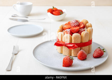 Charlotte, francese dessert con fragole, legato con un nastro rosso scarlatto, tazza di caffè, la ciotola con le fragole sul tavolo bianco. Foto Stock