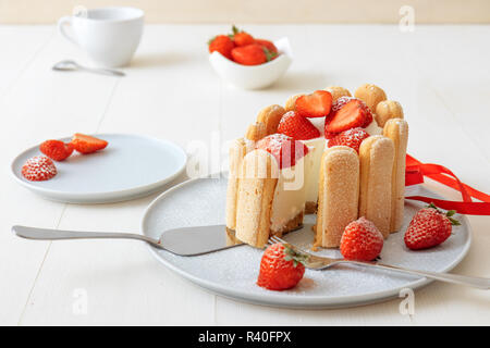 Charlotte, francese dessert con fragole, legato con un nastro rosso scarlatto, tazza di caffè, la ciotola con le fragole sul tavolo bianco. Foto Stock