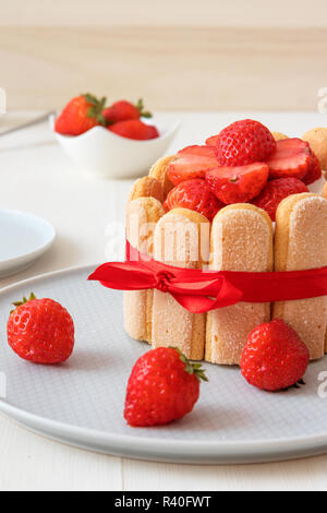 Charlotte, francese dessert con fragole, legato con un nastro rosso scarlatto, tazza di caffè, la ciotola con le fragole sul tavolo bianco. Foto Stock