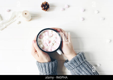 Composizione di natale. Donna con le mani in mano in maglione holding tazza di cioccolata calda. Marshmallows, pigne e nastro di seta bianca sul tavolo di legno dello sfondo. Winter Colazione concetto. Appartamento laico, vista dall'alto. Foto Stock