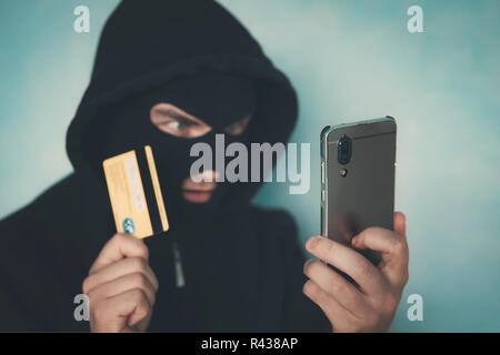 Close up di un uomo in maschera di rapina e cappa tenendo la carta di credito e guardando lo schermo dello smartphone. Penale maschio predispone un affare finanziario wit Foto Stock