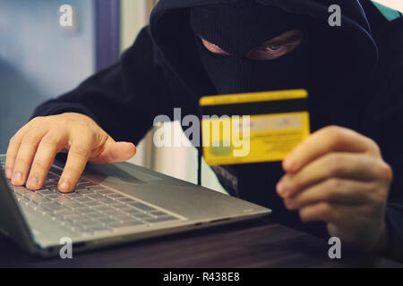 Uomo in maschera rapinatore utilizza internet, conto corrente bancario e di credito. Attacco di phishing da maschio con faccia nascosta. Il pirata informatico entra il furto di dati finanziari. C Foto Stock