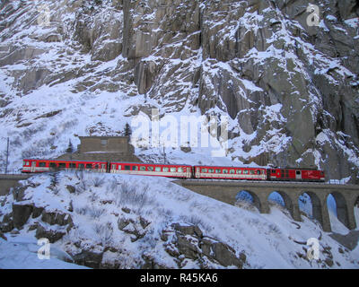 Montagna rocciosa scogliera con red Matterhorn Gotthard Bahn treno ferroviario guidando attraverso il bianco delle alpi svizzere attraversando un ponte di arco coperto di neve e ghiaccio Foto Stock