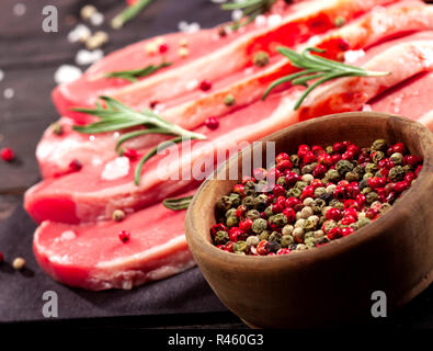 La carne cruda. Materie bistecca su un tagliere con rosmarino e spezie su uno sfondo nero. Foto Stock