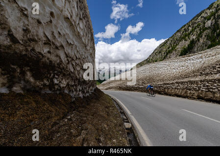 Ciclista scorre tra alti muri di neve lungo la strada a solo aprire Nufenen pass montagna nelle Alpi svizzere in giugno Foto Stock