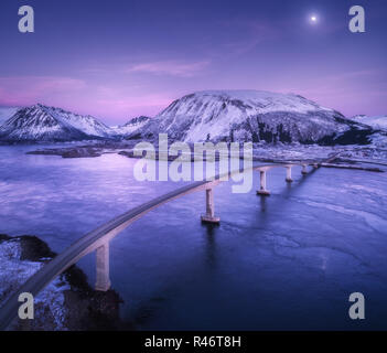 Vista aerea del ponte, montagne coperte di neve, viola il cielo con le nuvole e la luna, riflesso nell'acqua. Paesaggio notturno con bridge, rocce innevate, blu se Foto Stock