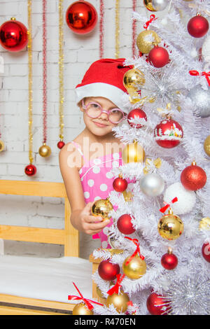 La ragazza in bicchieri arrotondati di rosso e un cappello di Natale si blocca le sfere su un nuovo nevoso anni albero di Natale Foto Stock