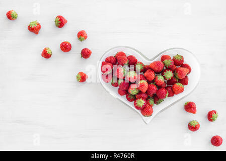 Le fragole in un cuore-ciotola sagomata, sul bianco sullo sfondo di legno. Foto Stock