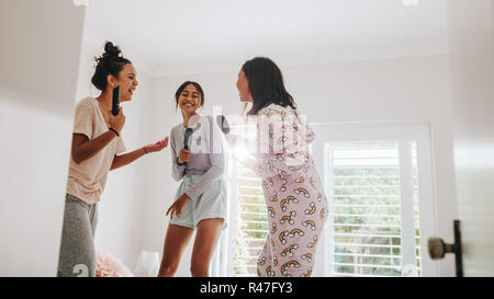 Tre ragazze in piedi sul letto azienda spazzole per capelli e telecomando a cantare in stile karaoke. Le fanciulle felici a sleepover godendo di cantare insieme. Foto Stock