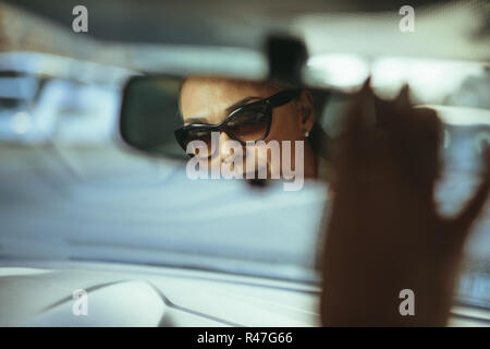 Senior driver donna regolare vista posteriore specchio auto. La riflessione a specchio retrovisore di senior donna che indossa gli occhiali da sole. Foto Stock