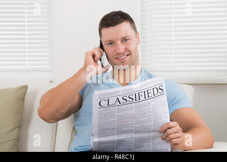 Uomo che legge il giornale mentre si utilizza il telefono cellulare a casa Foto Stock