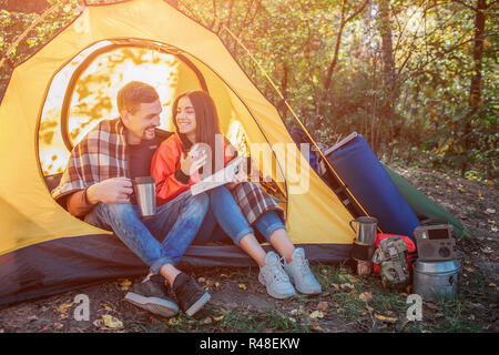 Giovani peoaple seduto in tenda e guardare a ogni altro. Essa detiene il libro aperto mentre il ragazzo ha thermos in mani. Essa detiene thermocup. Vi zaino con apparecchiature a tenda. Foto Stock
