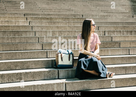 Bella ragazza turistico o studente in occhiali da sole e con uno zaino siede su per le scale e si appoggia. Stile di vita Foto Stock