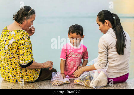 Due donne e bambino con spuntini al tramonto sul Mekong Riverfront Promenade; Preah Sisowath Quay, centro della città di Phnom Penh, Cambogia Foto Stock