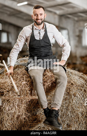 Ritratto di un bel contadino nel grembiule seduti sul fieno nella stalla di fattoria di capra Foto Stock