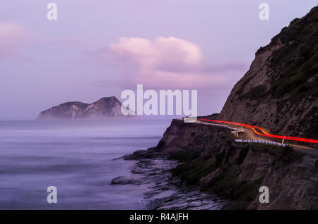 Luci rosse di una vettura guida su un telecomando East Cape strada costiera con telecomando isola, nebbia e ripida scogliera rendendo il paesaggio Foto Stock