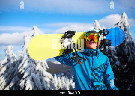 Snowboarder sorridente in posa che porta snowboard sulla spalla alla  stazione sciistica vicino a forest prima di backcountry freeride e  indossando occhiali riflettenti e colorati di moda vestito. Moderne  attrezzature di snowboard