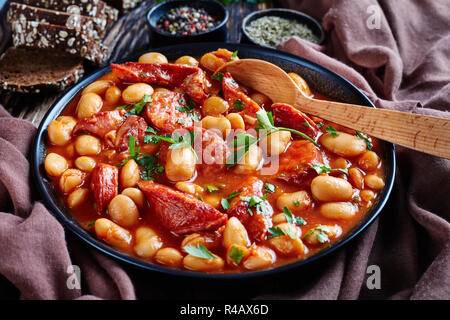 Stufato di fagioli con salsicce, erbe aromatiche e spezie in salsa di pomodoro in una ciotola nera con cucchiaio, su una tavola in legno rustico con panno marrone e pane, close-up Foto Stock