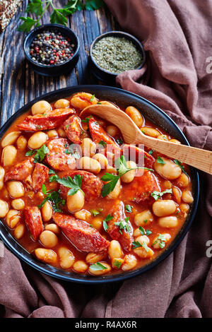 Stufato di fagioli con salsicce, erbe aromatiche e spezie in salsa di pomodoro in una ciotola nera con cucchiaio, su una tavola in legno rustico con panno marrone, fasolka bretonsku po Foto Stock