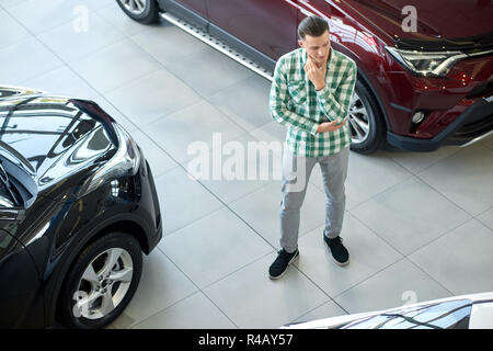 Giovane uomo in piedi tra nero e rosso scuro automobili. Ragazzo in camicia a scacchi e pantaloni grigi pensieroso guardando sul lato. Bella bionda client scegliendo nuovo veicolo e tenendo una mano vicino a mento. Foto Stock