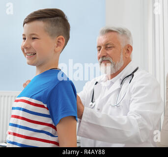 Medico guardando alla schiena del paziente ed esaminando la postura del ragazzino. Capretto felice sorridente, guardando lontano. Medico avente uno stetoscopio sul collo, indossa in bianco camice. Foto Stock