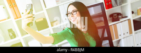 Una giovane ragazza con gli occhiali si siede in ufficio a tavola e scatta foto di se stessa sul telefono. Foto Stock