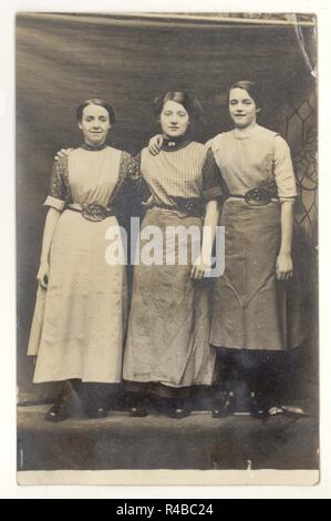 WW1 era studio ritratto di 3 ragazze, forse Lancashire lavoratori mulino di cotone che indossano zoccoli e grembiuli con gancio-come strumento e forbici nelle loro cinture appositamente progettato, circa 1915,1918, Regno Unito Foto Stock