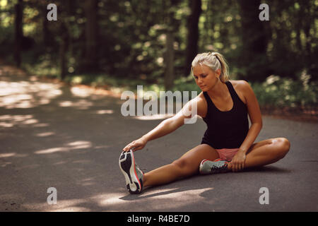 Montare la giovane donna bionda in sportswear seduto su una strada forestale stretching le gambe prima di una corsa Foto Stock