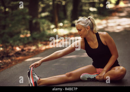 Montare la giovane donna bionda in sportswear stretching le gambe mentre è seduto sulla strada di un paese prima di una corsa Foto Stock