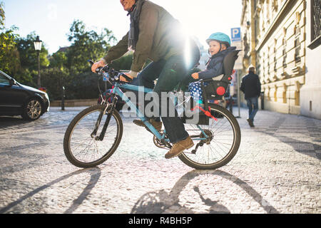 Un piccolo bimbo ragazzo seduto in sella per bicicletta con padre all'aperto in città. Foto Stock