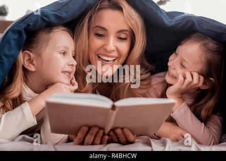 Storie emozionanti. Bella donna piacevole la lettura di un libro con i bambini mentre posa sul letto Foto Stock