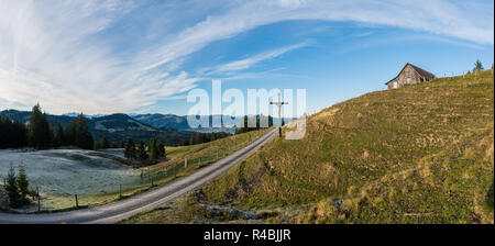 Aprire il paesaggio di campagna con prato erboso e la strada e la brina e il ghiaccio e la vecchia Croce di legno nel mezzo sotto un cielo espansiva e una capanna in legno fino a t Foto Stock