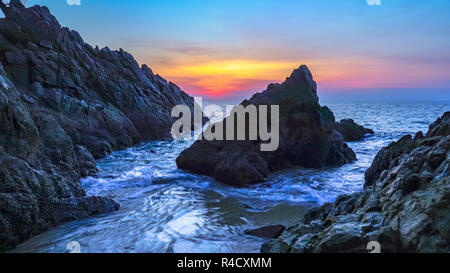 Le onde battono il grande cono roccia al centro di pietre cape durante il tramonto al Banana beach Phuket Foto Stock