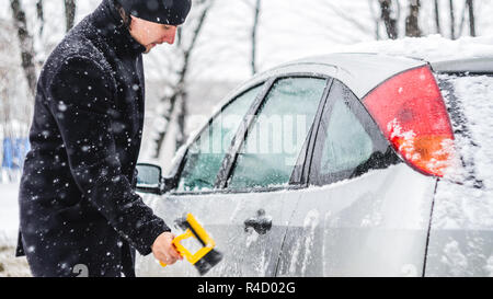 Giovane uomo in cappotto nero pulisce la sua vettura con il pennello di colore giallo durante la nevicata. Inverno condizioni atmosferiche avverse. Foto Stock