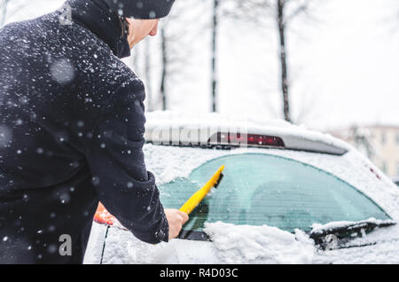 Giovane uomo in cappotto nero pulisce la sua vettura con il pennello di colore giallo durante la nevicata. Inverno condizioni atmosferiche avverse. Foto Stock