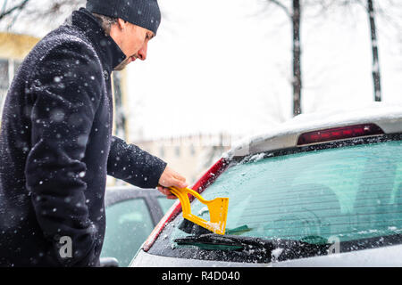 Giovane uomo in cappotto nero pulisce la sua vettura con il pennello di colore giallo durante la nevicata. Inverno condizioni atmosferiche avverse. Foto Stock