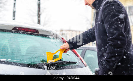 Giovane uomo in cappotto nero pulisce la sua vettura con il pennello di colore giallo durante la nevicata. Inverno condizioni atmosferiche avverse. Foto Stock