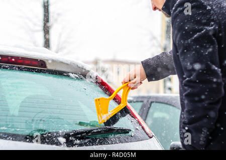 Giovane uomo in cappotto nero pulisce la sua vettura con il pennello di colore giallo durante la nevicata. Inverno condizioni atmosferiche avverse. Foto Stock