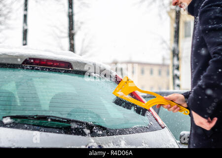 Giovane uomo in cappotto nero pulisce la sua vettura con il pennello di colore giallo durante la nevicata. Inverno condizioni atmosferiche avverse. Foto Stock