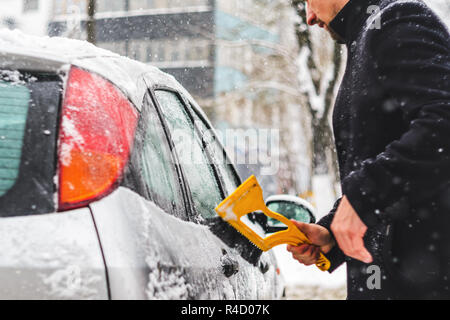 Giovane uomo in cappotto nero pulisce la sua vettura con il pennello di colore giallo durante la nevicata. Inverno condizioni atmosferiche avverse. Foto Stock