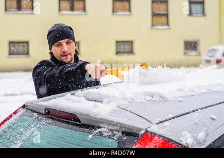 Giovane uomo in cappotto nero pulisce la sua vettura con il pennello di colore giallo durante la nevicata. Inverno condizioni atmosferiche avverse. Foto Stock