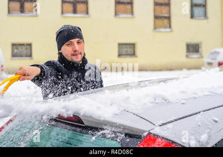 Giovane uomo in cappotto nero pulisce la sua vettura con il pennello di colore giallo durante la nevicata. Inverno condizioni atmosferiche avverse. Foto Stock