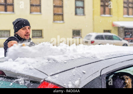 Giovane uomo in cappotto nero pulisce la sua vettura con il pennello di colore giallo durante la nevicata. Inverno condizioni atmosferiche avverse. Foto Stock