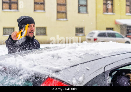 Giovane uomo in cappotto nero pulisce la sua vettura con il pennello di colore giallo durante la nevicata. Inverno condizioni atmosferiche avverse. Foto Stock